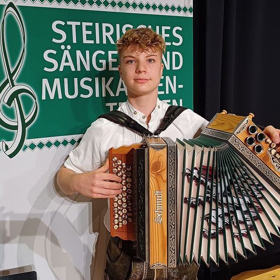 Ein junger Mann steht vor einem grünen Schild und hält ein Akkordeon. Das Schild trägt die Aufschrift 'Steirisches Sänger- und Musiker-Ensemble'.