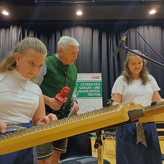 Zwei Frauen und ein Mann spielen eine Zither. Der Mann hält ein Mikrofon mit einem roten Logo. Die Frauen tragen weiße Oberteile, und der Mann trägt eine grüne Weste. Hinter ihnen steht ein Mikrofonstativ und ein Banner mit der Aufschrift 'Steirisches Sanger-und Musikanten-Treffen.'