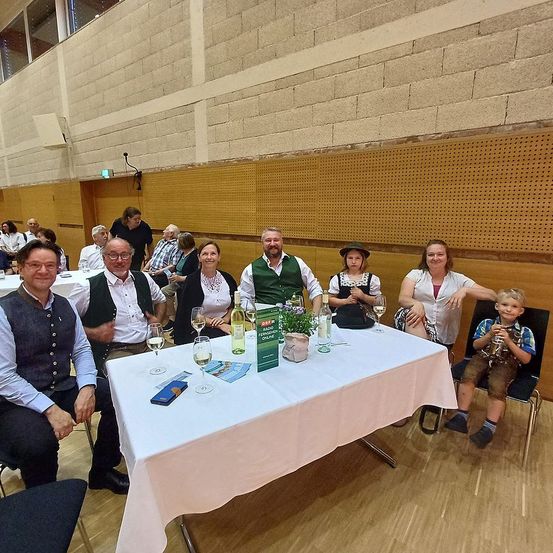 Eine Gruppe von Erwachsenen und einem Kind in traditioneller bayerischer Kleidung sitzen um einen Tisch mit Weingläsern und einem Buch. Sie befinden sich in einem Raum mit Holz- und Ziegelwänden.