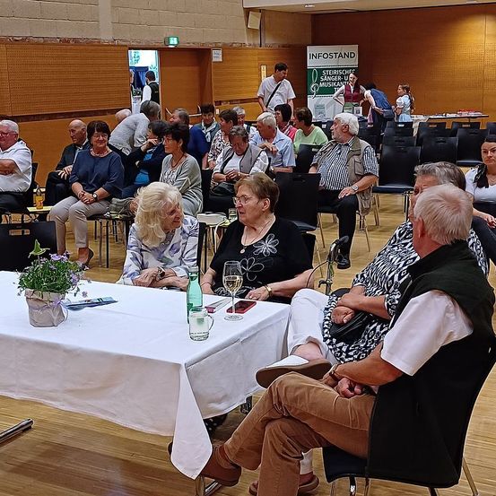Eine Gruppe älterer Menschen sitzt in einem Raum, wahrscheinlich einem Konferenzsaal. Sie tragen formelle Kleidung und einige von ihnen tragen Brillen. Ein Tisch mit einer Blumenvase und einer Flasche steht vor ihnen. Es gibt Stühle und einen Schreibtisch an der Seite. Ein Plakat ist an der Wand angebracht.