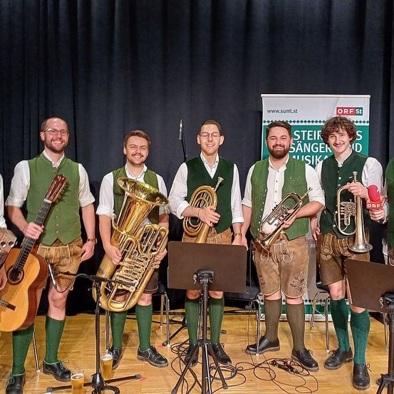 Fünf Männer in traditioneller grüner Kleidung und braunen Hosen stehen vor einem schwarzen Vorhang und halten Musikinstrumente. Sie lächeln für die Kamera. Hinter ihnen steht ein Notenständer auf dem Boden. Ein Poster mit dem Text 'Steirer Sänger Musik' ist an der Wand befestigt. Ein Stativ ist vor ihnen platziert. Ein Mikrofon ist auf dem Stativ befestigt.
