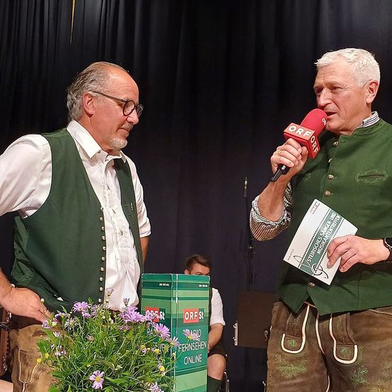 Zwei Männer auf der Bühne, einer hält ein Mikrofon und spricht, der andere steht neben einem Blumenstrauß. Der Mann mit dem Mikrofon trägt eine grüne Weste und hält ein Papier. Der Hintergrund ist schwarz.