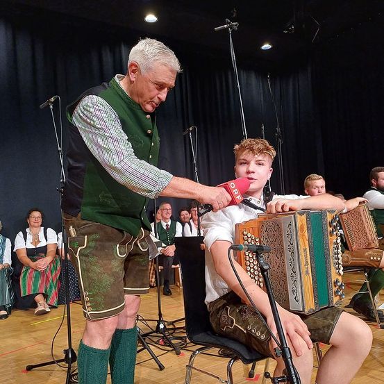 Ein älterer Mann mit Mikrofon interviewt einen jungen Mann, der auf der Bühne ein Akkordeon spielt, vor einem sitzenden Publikum.