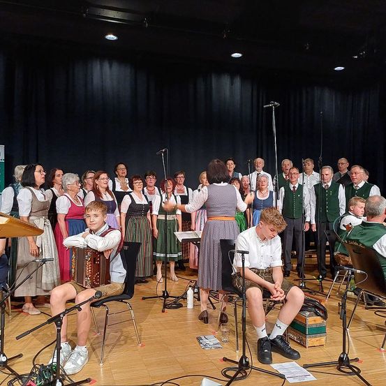 Ein Chor und eine Band sind auf der Bühne mit Mikrofonen. Zwei sitzende Jungen spielen Akkordeon und Klavier. Der Chor steht und singt. Musikinstrumente und Ausrüstung sind vorhanden.