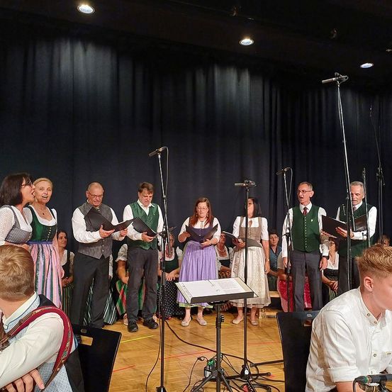 Ein Chor aus Erwachsenen in traditioneller bayerischer Kleidung tritt auf der Bühne mit Mikrofonen auf. Das Publikum sitzt im Hintergrund.