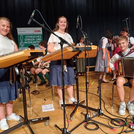 Eine Gruppe junger Menschen spielt auf der Bühne Musikinstrumente. Zwei Mädchen spielen Zither und ein Junge Akkordeon. Die Szene ist lebhaft mit Zuschauern im Hintergrund.