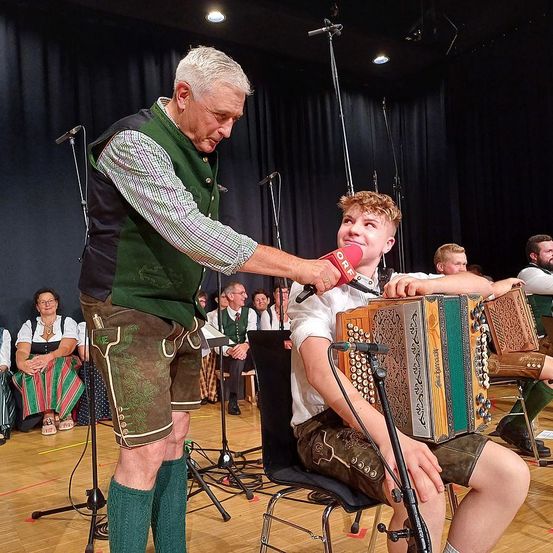 Ein älterer Mann interviewt einen jungen Jungen, der ein Akkordeon spielt, während er traditionelle bayerische Kleidung trägt. Sie befinden sich in einem Studio mit einem Mikrofon und Publikum hinter ihnen.