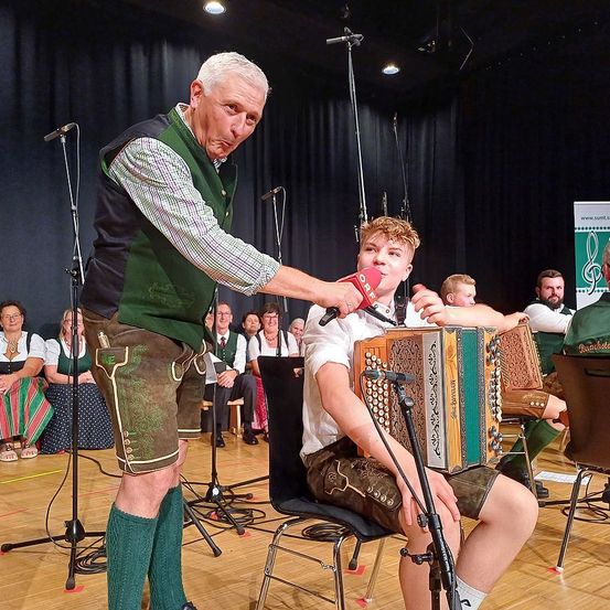 Ein älterer Mann steht auf der Bühne und hält ein Mikrofon an einen jungen Jungen, der ein Akkordeon spielt. Der Junge sitzt auf einem Stuhl vor einem Mikrofon. Dahinter sitzt eine Gruppe von Menschen und schaut zu.