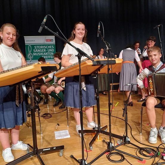 Zwei junge Frauen spielen eine Zither mit Mikrofonen auf der Bühne, während ein Junge ein Akkordeon spielt. Ein Schild mit der Aufschrift 'Steirisches Sänger-und Musikanten-Treffen' ist im Hintergrund. Andere Musiker spielen Instrumente und singen.