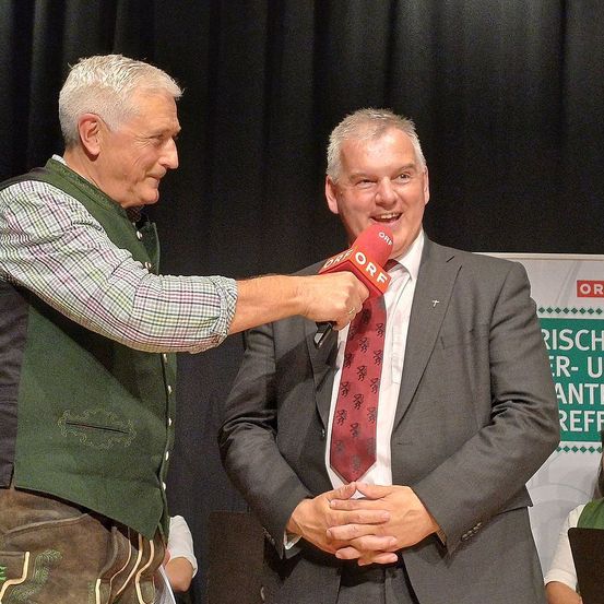 Zwei Männer auf einer Bühne, einer hält ein Mikrofon und der andere lächelt. Der Mann mit dem Mikrofon trägt eine grüne Weste und ein kariertes Hemd, während der lächelnde Mann einen Anzug und eine Krawatte trägt. Hinter ihnen ist ein schwarzer Hintergrund mit einer Werbung.