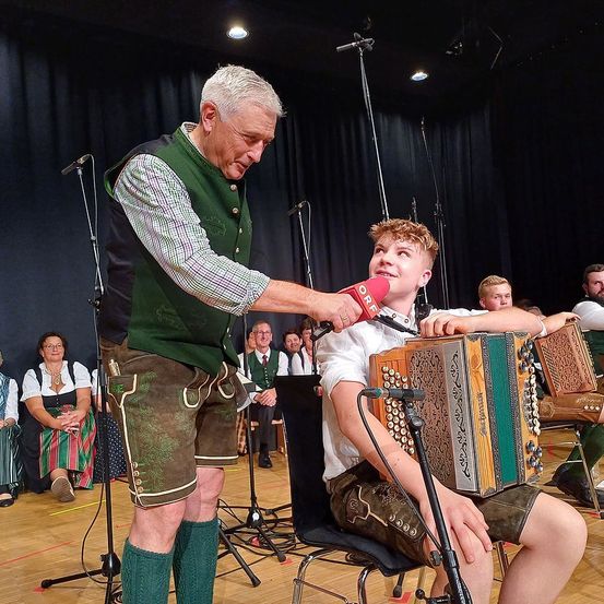 Ein älterer Mann spricht in ein Mikrofon, während er es vor einem jungen Jungen hält, der ein Akkordeon spielt. Dahinter sitzen Menschen auf Stühlen und beobachten die Aufführung.