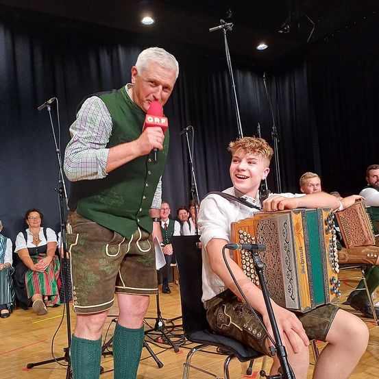 Ein Mann mit einem Mikrofon steht neben einem jungen Mann, der Akkordeon spielt. Sie sind beide auf einer Bühne mit schwarzem Vorhang. Andere Leute sitzen im Hintergrund.