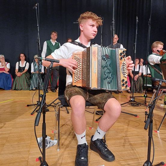 Ein junger Mann spielt Akkordeon während einer Aufführung mit anderen Musikern auf der Bühne. Hinter ihm sitzen und stehen Menschen und beobachten.