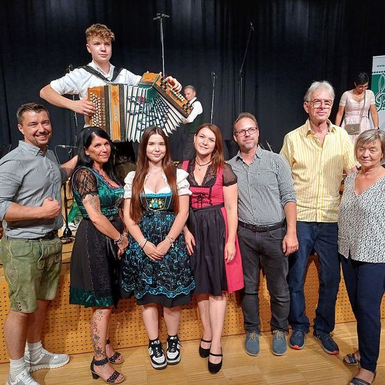 Eine Gruppe von Menschen auf der Bühne, darunter ein Mann mit einem Akkordeon, stehen zusammen mit einem Hintergrund aus schwarzen Vorhängen und Mikrofonen. Einige sind in traditioneller Kleidung gekleidet.