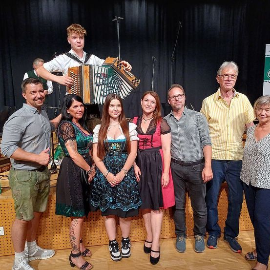 Eine Gruppe von Menschen posiert für ein Foto, zwei Frauen in traditioneller Kleidung, ein Mann hält ein Akkordeon, und andere in lässiger Kleidung. Sie stehen auf einer Bühne mit schwarzem Hintergrund.