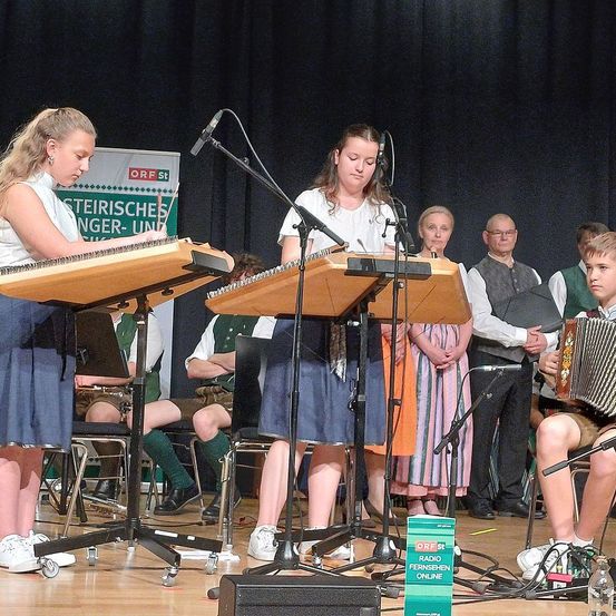 Zwei Mädchen spielen Holzxylophone auf einer Bühne mit schwarzem Hintergrund. Ein Junge spielt Akkordeon auf der rechten Seite. Dahinter stehen mehrere Menschen und schauen zu.