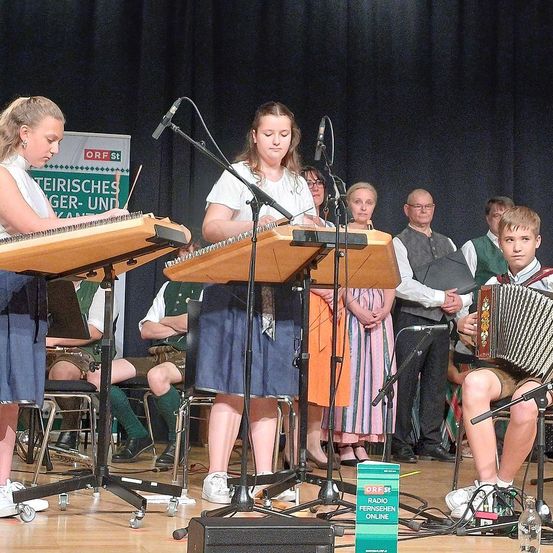 Zwei Mädchen spielen Holzxylophone auf der Bühne, während ein Junge Akkordeon spielt. Hinter ihnen sitzen und stehen mehrere Personen. Die Bühne hat einen Hintergrund mit einem Banner und einem Mikrofonständer.
