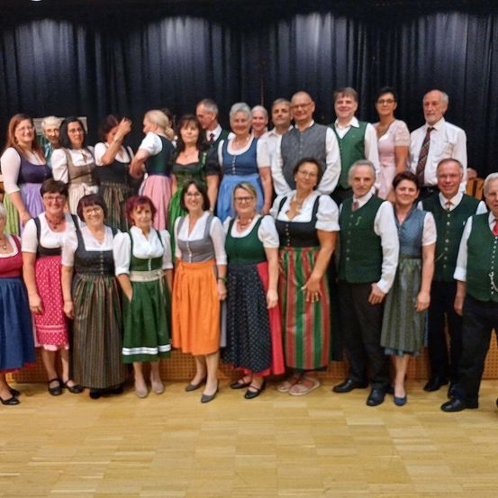 Eine Gruppe von Menschen, wahrscheinlich ein Chor, posiert für ein Foto auf einer Bühne mit Holzboden. Sie tragen traditionelle Kostüme.