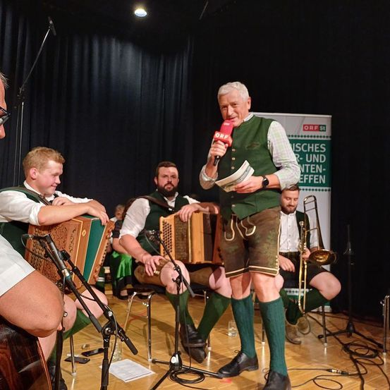 Eine Gruppe von Musikern tritt auf der Bühne auf. Der Mann in der Mitte hält ein Mikrofon und scheint zu sprechen oder zu singen. Ein anderer Mann auf der linken Seite spielt Akkordeon, während ein anderer Mann auf der rechten Seite Posaune spielt. Am Rücken ist ein Banner mit Text angebracht.