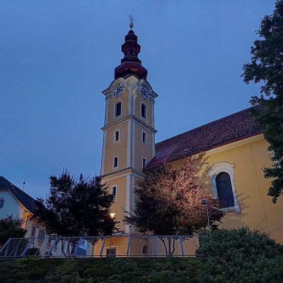 Ein hoher Kirchturm mit einer Uhr, umgeben von Bäumen und Büschen. Der Turm ist beige mit einem roten Dach, und die Uhr ist auf zwei Seiten sichtbar.