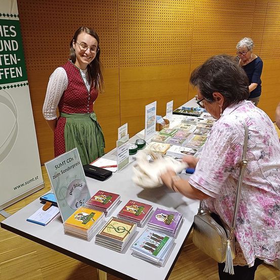 Eine Frau in traditioneller Kleidung steht hinter einem Tisch mit CDs. Eine andere Frau schaut sich die CDs an. Ein Banner mit dem Text 'Sound und Licht' ist sichtbar.