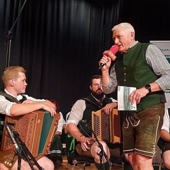 Ein Mann, der ein Mikrofon hält, spricht in dieses, während er vor zwei Personen steht, die Akkordeon spielen. Hinter ihnen ist ein schwarzer Vorhang zu sehen.