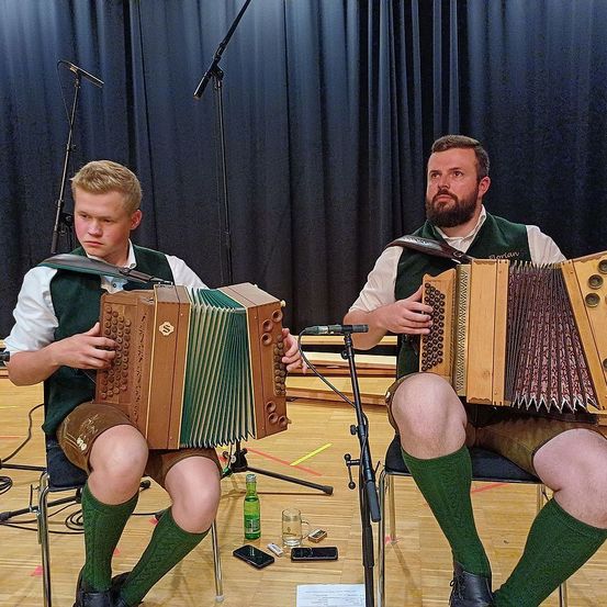 Zwei Männer in traditioneller Kleidung spielen Akkordeons auf einer Bühne. Einer sitzt mit einem Mikrofon vor sich, während der andere neben ihm sitzt. Dahinter ist ein schwarzer Vorhang gezogen.