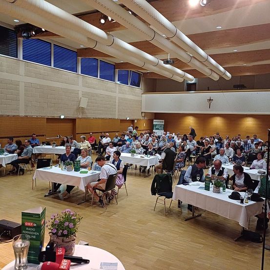Ein Saal mit vielen Menschen, die an Tischen sitzen, mit weißen Tischdecken und Weingläsern. Ein Mikrofon und Blumen sind auf der linken Seite. Eine Projektionslampe ist an der Decke montiert.