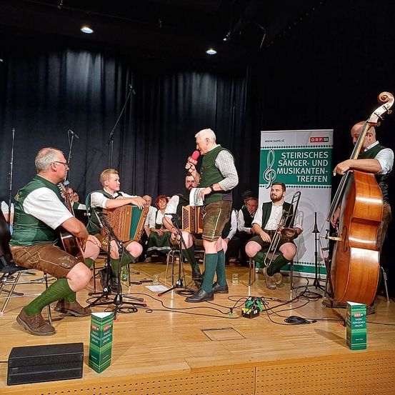 Eine Gruppe Musiker tritt auf der Bühne auf. Sie sind in traditioneller Kleidung gekleidet. Ein Banner im Hintergrund lautet Steirisches Sänger-und Musikanten-Treffen.