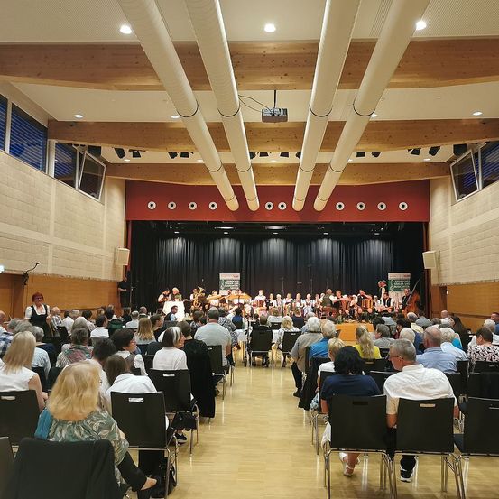 Eine Gruppe von Menschen sitzt in Stühlen in einem Saal. Eine Band spielt auf der Bühne vor ihnen. Es gibt Lautsprecher und einen Projektor auf der Bühne. Die Decke hat Holzbalken und einen Projektor. Es gibt Fenster an der Wand.