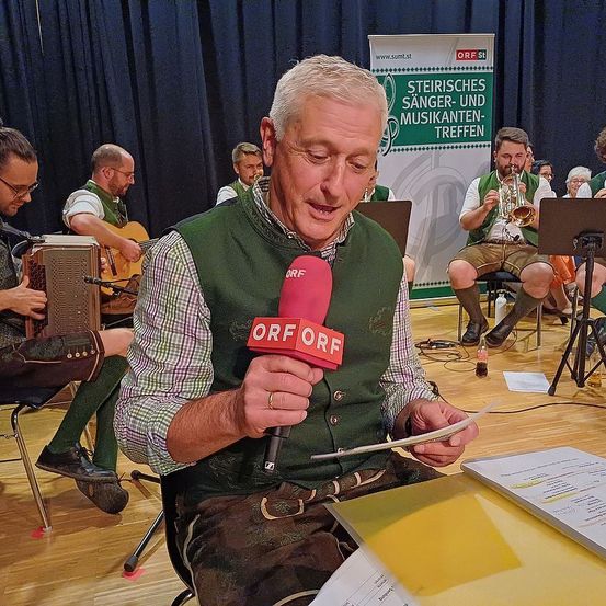 Ein Mann hält ein Mikrofon mit ORF darauf. Er trägt eine grüne Weste. Hinter ihm spielt eine Gruppe von Menschen Musikinstrumente. Im Hintergrund befindet sich ein Banner mit Text.