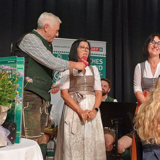 Eine Frau in einem traditionellen deutschen Kleid steht auf der Bühne und wird von einem Mann mit einem Mikrofon interviewt. Hinter ihnen befindet sich ein Banner mit grünem Text, und ein Publikum ist teilweise sichtbar.