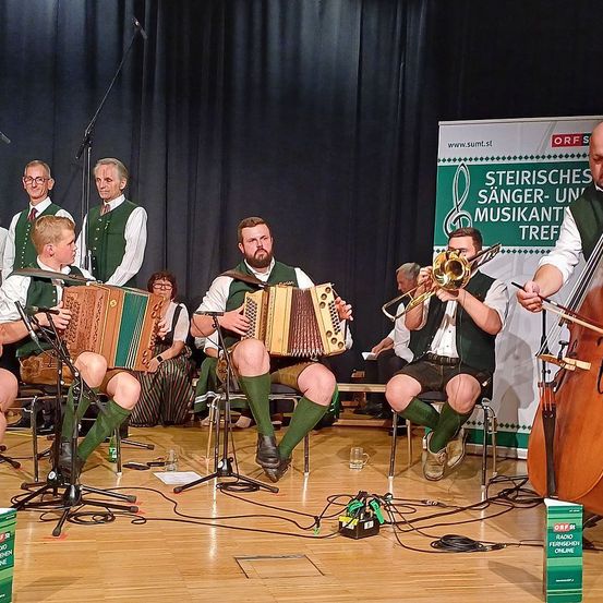 Eine Gruppe von Musikern spielt auf der Bühne und verwendet Instrumente wie Akkordeon und Cello. Sie sitzen auf Stühlen mit einem Banner hinter ihnen, eines mit der Aufschrift 'Steirische Sanger-Un Musikant'. Ein Mikrofon und Ständer sind vor ihnen aufgestellt.