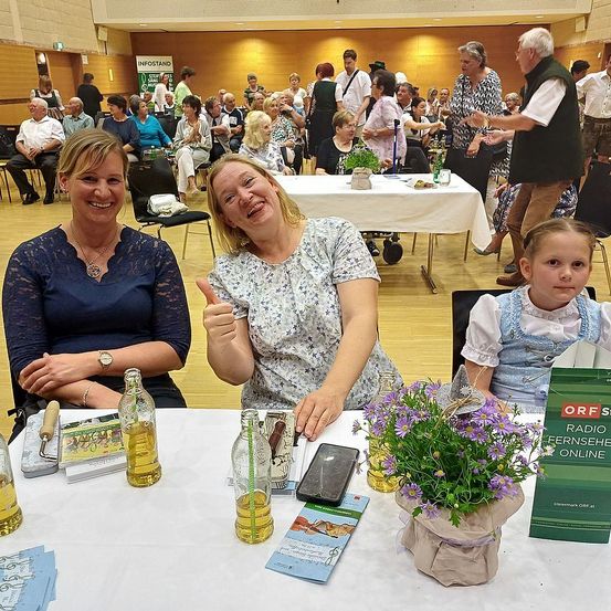 Drei Frauen und ein junges Mädchen sitzen an einem Tisch mit Blumen, Flaschen und Büchern. Hinter ihnen sitzen und stehen eine Gruppe von Menschen in einem Saal.