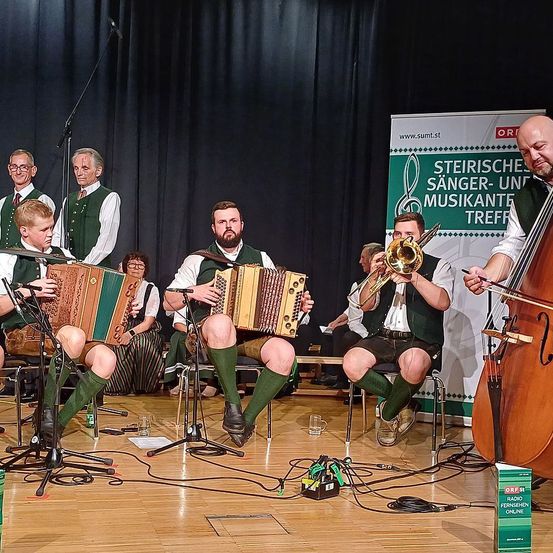 Eine Gruppe von Musikern spielt Instrumente auf der Bühne, darunter Akkordeons, eine Trompete und eine Geige. Dahinter befindet sich ein Banner mit Text und Logos. Die Umgebung ist drinnen mit einem Holzboden.