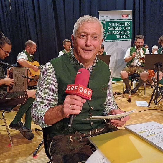 Ein Mann mit einem Mikrofon hält ein Blatt Papier in den Händen. Hinter ihm spielen Musiker verschiedene Instrumente auf der Bühne. Das Banner dahinter lautet 'Steirisches Sänger-und Musikertreffen'.