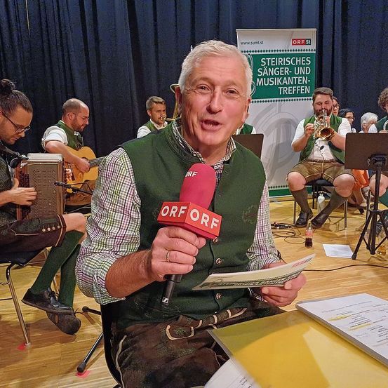 Ein Mann mit einem Mikrofon mit ORF darauf hält es an und spricht hinein, während er auf einem Stuhl sitzt. Dahinter spielt eine Band auf der Bühne, einschließlich Akkordeon, Gitarre und Trompete. Ein Banner im Hintergrund lautet 'Steirisches Sänger-und Musikanten-Treffen'.