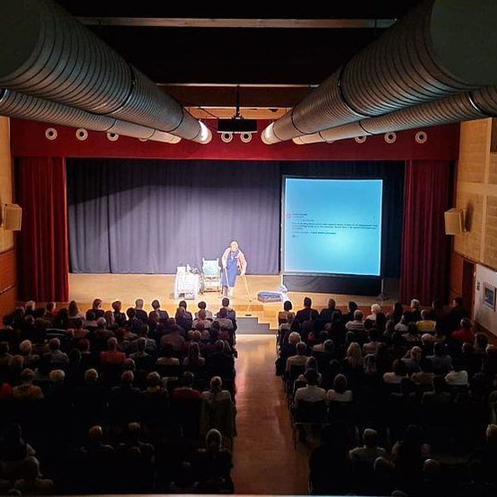 Ein großer Innenraum mit Bühne, Leinwand und Lautsprecher an der Wand. Eine Person steht auf der Bühne mit einem Beamer. Das Publikum sitzt auf Stühlen vor der Bühne.