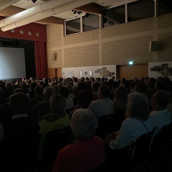 Ein volles Auditorium mit einer großen Leinwand auf der Bühne, sitzende Menschen, rote Vorhänge und mehrere gerahmte Bilder an der Wand.