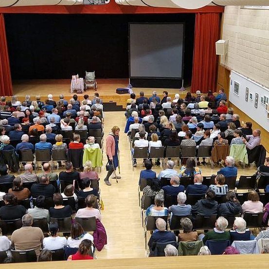 Ein überdachter Saal mit einem Publikum, das auf Stühlen sitzt. Eine große Leinwand und eine Bühne sind sichtbar. Mehrere Personen stehen auf der Bühne.