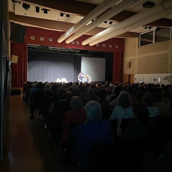 Ein Auditorium mit Bühne und Publikum. Zwei Personen stehen auf der Bühne. Der Raum hat weiße Wände, einen roten Vorhang und eine Projektionsleinwand.