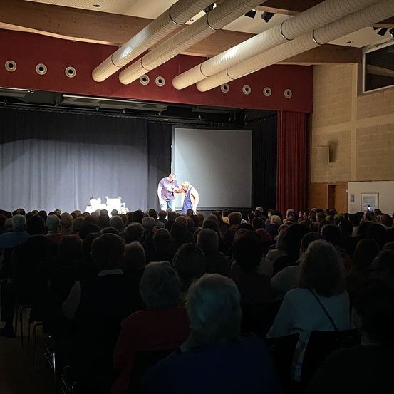 Eine Theaterbühne mit zwei Personen vor einer Leinwand, ein Publikum schaut von den Stühlen unten zu.