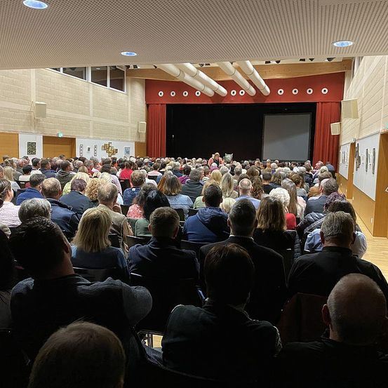 Ein volles Auditorium mit vielen sitzenden Personen. Im Vordergrund ist eine Bühne mit Leinwand und Vorhängen zu sehen.