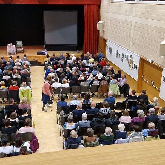 Eine große Gruppe von Menschen sitzt in einem Auditorium auf Stühlen und schaut sich eine Präsentation auf einer großen Leinwand an. Der Raum hat Holzböden und weiße Wände mit Bildern und Gemälden.