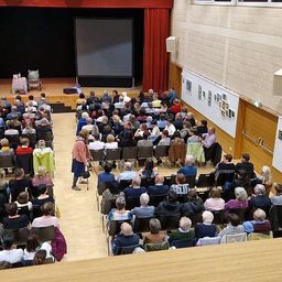 Eine große Gruppe von Menschen sitzt in einem Auditorium auf Stühlen und schaut sich eine Präsentation auf einer großen Leinwand an. Der Raum hat Holzböden und weiße Wände mit Bildern und Gemälden.