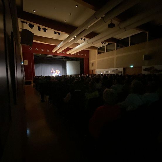 Ein Innentheater mit roter Bühnenvorhang, einer Bühne und Menschen im Dunkeln sitzend. Zwei Personen stehen auf der Bühne, beleuchtet von einem Scheinwerfer.