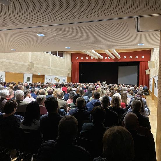 Ein großes Publikum sitzt in einem modernen Auditorium mit einer Bühne mit Projektorleinwand und roten Vorhängen, mit Reihen von Menschen, die der Bühne zugewandt sind, einige mit ihrer Aufmerksamkeit auf den Bildschirm gerichtet.