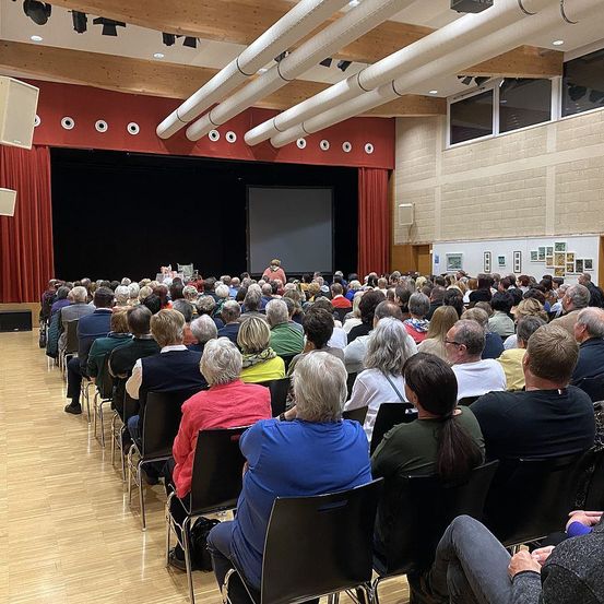 Ein Raum mit Holzböden ist mit Menschen gefüllt, die in Reihen sitzen. Eine Bühne mit einer Projektionsleinwand befindet sich vorne, und rote Vorhänge sind darum herum. Eine Frau steht auf der Bühne und hält wahrscheinlich eine Präsentation ab.