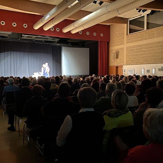 Ein Indoor-Saal mit einer großen Leinwand und zwei Personen, die auf der Bühne auftreten, während eine Menschenmenge in Stühlen sitzt und zuschaut.