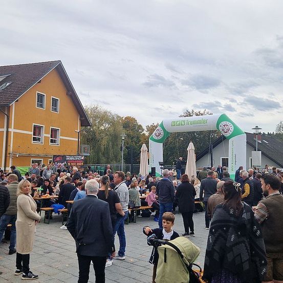 Eine Menschenmenge versammelt sich draußen um Tische unter einem Bogen, einige tragen Hüte und Schuhe, mit einem gelben Gebäude und Bäumen im Hintergrund.